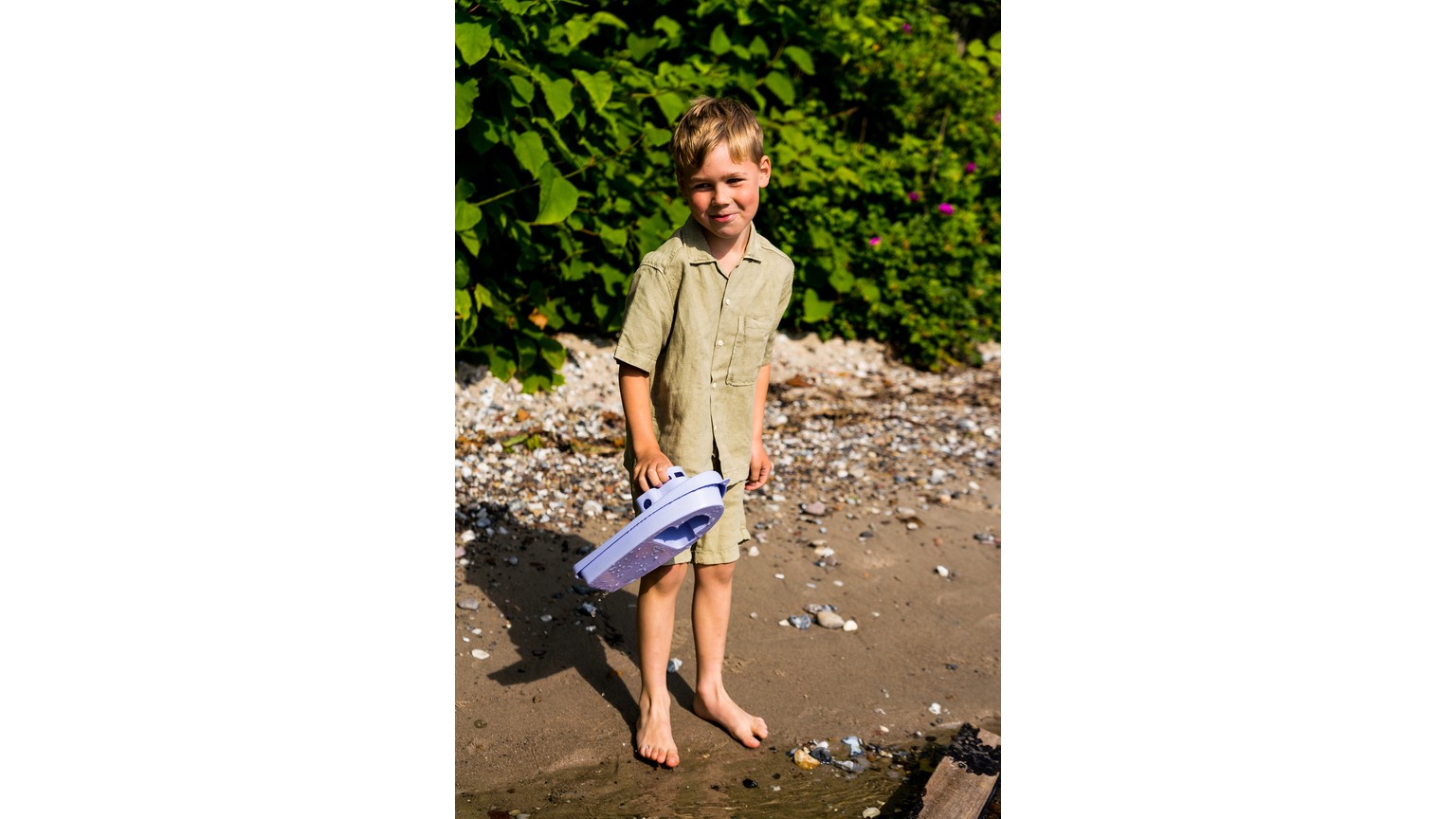 Barn på strand med legetøjsbåd i hånden i solskinsvejr med grøn bevoksning i baggrunden.